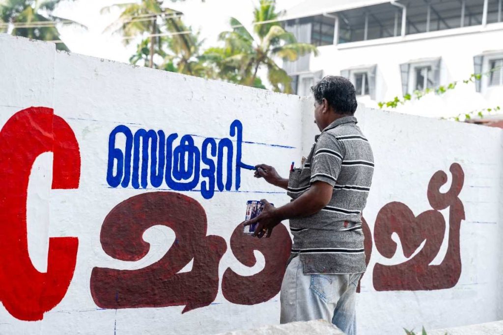 Wall painting artist painting Malayalam letters on a white wall.