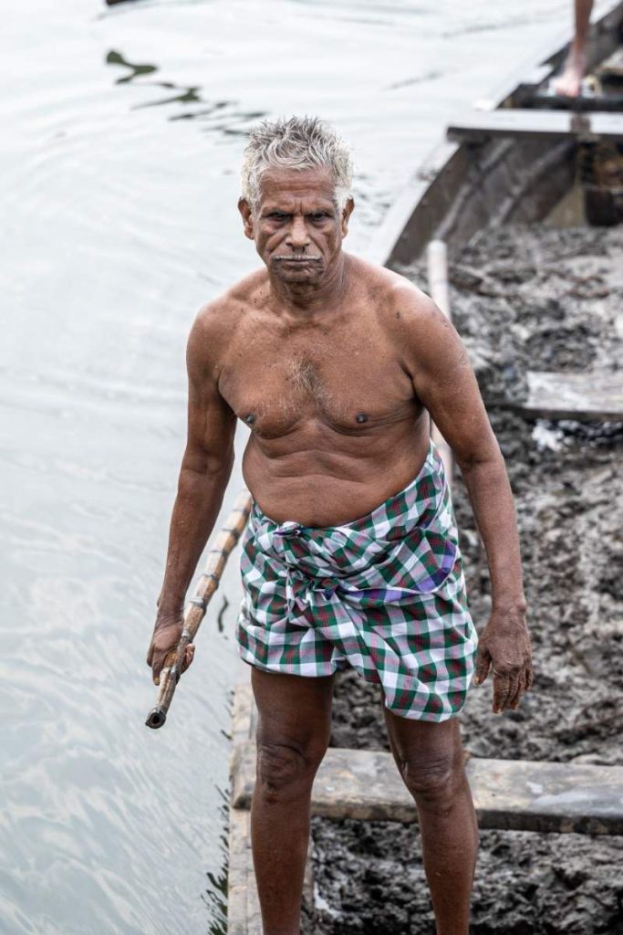 Elderly clay laden man in checkered lungi on a canoe.