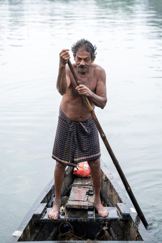 Smiling elderly man creating tradition in dark lungi rowing canoe.