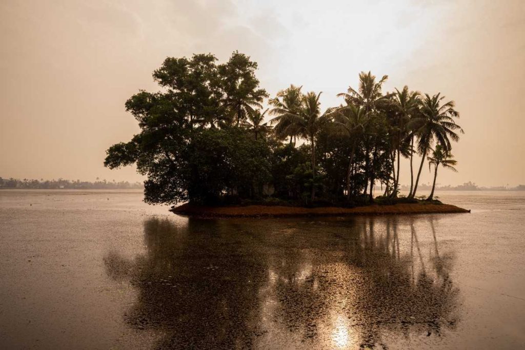 Sun lit skies with a small island with trees reflecting on calm water at sunrise.