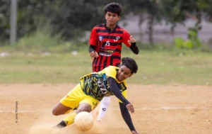 Footballer in yellow sliding to win the ball in the dust, sports photograph by John Thomas, Creative Hut Academy